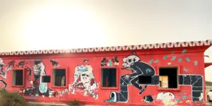 קיר אדום בהיר של בניין עם אמנות גרפיטי סוריאליסטית מציג דמויות מופשטות של אדם ויצור. היצירה כוללת שלד בחליפה וכסף מפוזר, עם חלונות משולבים לאורך הקיר - אחד המקומות הייחודיים מומלצים לטיול למטיילים בישראל.