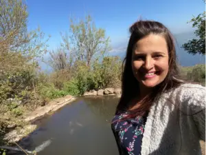 אישה מחייכת לסלפי ליד בריכה טבעית שלווה, עטופה בעצים ושיחים שופעים. השמיים כחולים צלולים, ממסגרים גבעות רחוקות ומים שלווים - מקום מושלם לטיולים בצפון הארץ המופיעים בבלוג טיולים בארץ שלה.