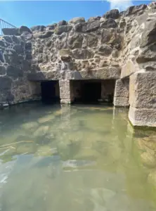 מבנה אבן עם פתחים מקושתים, שקוע בחלקו במים צלולים. פני השטח משקפים את השמיים, והפרזול מחוספס ועם מרקם. אור השמש מאיר חלקים מהסצנה הזו, מציג את הפיתוי העתיק שלה - פנינה אמיתית למטיילים בישראל המחפשים פלאים נסתרים במסעם.