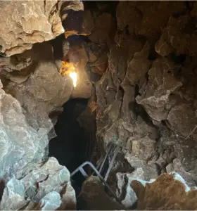 פתח מערה צר עם קירות סלעיים ומחוספסים מוביל כלפי מטה ומציע הצצה הרפתקנית לחוויות בטבע בישראל. סולם קטן נראה חלקית בתחתית, ואור קלוש מלמעלה מאיר את משטחי הסלע המרקמים, ומזמין מגלי טיולים בצפון הארץ.
