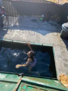 אדם שוחה במיכל מים מלבני חיצוני. מכסה המתכת הירוק פתוח חלקית. הקרקע מרוצפת, עם נעליים וקסדה בקרבת מקום. אור השמש מטיל צללים ברחבי האזור, לוכד את המהות של מטיילים בישראל במהלך יום ציורי של חקר והרפתקאות.