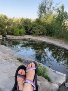 יושבים ליד בריכה קטנה עם רגליים נראות בסנדלים סגולים על שביל אבן, אפשר באמת להעריך את חוויות בטבע בישראל. הבריכה מוקפת בעשב גבוה ושיחים תחת שמיים כחולים צלולים, ומציעה רגעים שלווים המושלמים לבלוג טיולים בארץ.