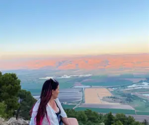 אדם עם שיער ארוך כהה וצעיף לבן יושב על גבעה ירוקה, טובל בנוף העצום של שדות והרים מרוחקים תחת שמיים כחולים צלולים בשקיעה - באמת אחד המקומות מומלצים לטיול למחפשים חוויות בטבע בישראל עוצרות נשימה.