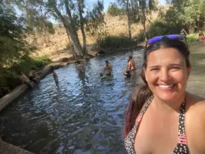 אישה מחייכת לסלפי מול בריכה טבעית קטנה שבה אנשים שונים שוחים ונרגעים. עצים וגבעות יבשות ודשא משמשות כרקע ליום שמש זה, ומציעות חוויות בטבע בישראל אמיתיות הלוכדות את המהות של מטיילים בישראל.