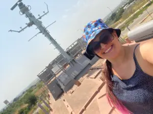 חייכן עם משקפי שמש וכובע דלי צבעוני מצלם סלפי בתוך הקסם של חוויות בטבע בישראל. ברקע, מבנה גבוה עם אנטנות משלים את הנוף הנופי של כבישים וירק מתחת.