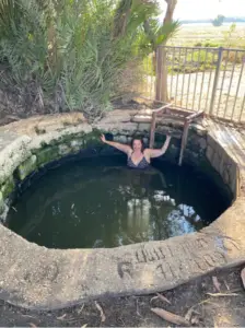 אדם בבגד ים מנוקד מחייך ונרגע בבריכת אבן קטנה ועגולה בינות לצמחייה עבותה. יום שמשי מאיר את הסצנה כשהם נהנים מהקסם של הטבע - חוויה הבולטת בין המקומות המומלצים לבקר בהם במטיילים בישראל.