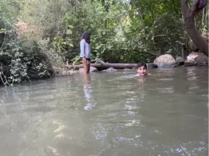 ילד שוחה בנחל מיוער, מחייך למצלמה, בעוד אדם עומד בסמוך על שפת הנחל בחולצה ארוכת שרוולים ומכנסיים קצרים. עצים ושיחים ירוקים שופעים מקיפים את המים, מציגים חוויות בטבע בישראל המדגישות את המקומות מומלצים לטיול.