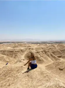 אדם לבוש בגופייה לבנה ומכנסיים קצרים כחולים יושב על גבעה סלעית ונהנה מאחד המקומות המומלצים לטיול. הנוף צחיח ונרחב תחת שמיים כחולים צלולים, מראה הרים רחוקים ואופק שטוח, האופייניים להרפתקאות בצפון ישראל.