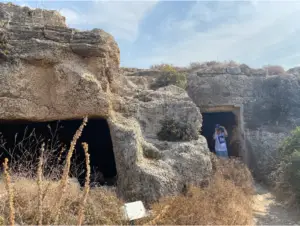 אדם עומד בפתח מערה חצובה בצלע גבעה סלעית מתחת לשמים כחולים צלולים. האזור, המופיע בבלוג טיולים רבים בארץ, מוקף בצמחייה יבשה ומתהדר בשלט קטן ליד המערה, מה שהופך אותו לאחד האטרקציות המסקרנות לאלו שמחפשים טיולים בצפון הארץ.