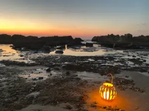 פנס זוהר יושב על חוף סלעי בשעת בין ערביים, אחד ממקומות מומלצים לטיול למי שמטייל בחוויות בטבע בישראל. השמיים הם שיפוע של כתום עד סגול כשהשמש שוקעת ברקע, עם בריכות מים המשקפות את האופק התוסס בין הסלעים על קו החוף.
