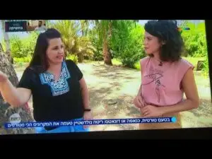 שתי נשים בחוץ בגן, חולקות את חוויותיהן של חוויות בטבע בישראל. האישה משמאל מחווה בידה, לובשת חולצה שחורה עם דוגמה לבנה. האישה מימין, בחולצה ורודה, מקשיבה בדריכות. טקסט בעברית נראה בתחתית.