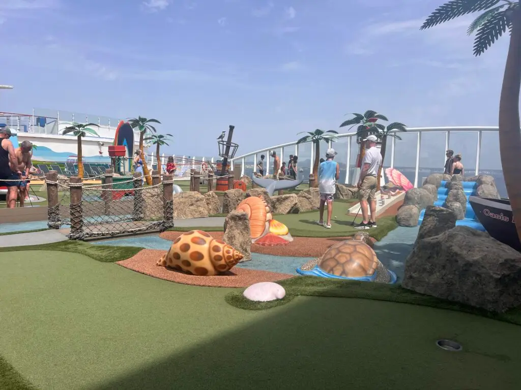 אנשים משחקים מיני גולף על סיפון ספינה מוקף עצי דקל מלאכותיים, צדפים דקורטיביים גדולים וסלעים תחת שמיים בהירים ומעוננים חלקית. גשר חבלים ונופי אוקיינוס נראים ברקע.