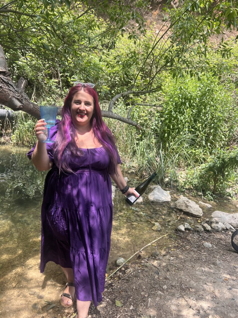 אישה עם שיער סגול ארוך ושמלה סגולה מחזיקה משקה ובקבוק בעודה עומדת ליד נחל באזור מיוער ביום שמש.