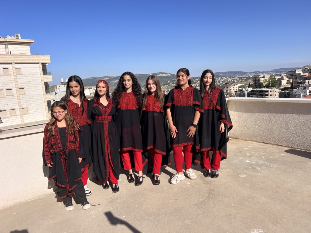 שבע בנות עומדות על גג לבושות בשמלות רקומות מסורתיות בשחור ואדום ומכנסיים אדומים, מחייכות כשברקע נשקפים שמיים כחולים בהירים ונוף עירוני.