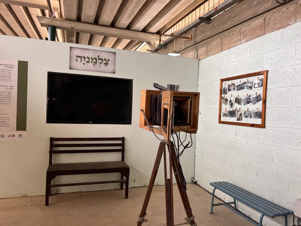 מצלמה וינטג' מעץ על חצובה ניצבת מול ספסל וטלוויזיה התלויה על הקיר. בקרבת מקום, קולאז' ממוסגר של תמונות בשחור-לבן תלוי על קיר לבנים לבן בחדר מואר היטב עם קורות חשופות.
