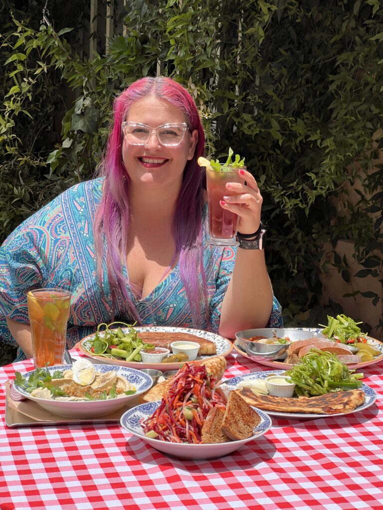 אישה מחייכת עם שיער ארוך ורוד וסגול אוחזת במשקה ליד שולחן חיצוני מכוסה במפה משובצת אדומה ולבנה, מוקפת בצלחות של אוכל וסלטים. עלווה ירוקה נראית ברקע.