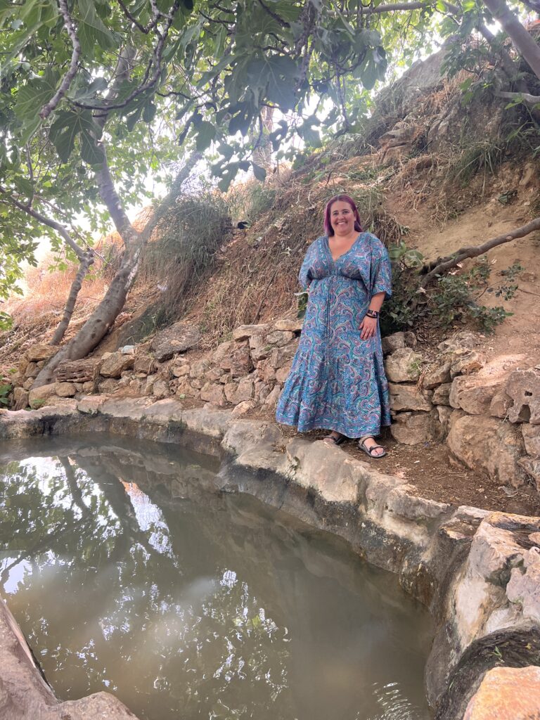 אישה בשמלה כחולה עם דוגמאות עומדת על מדף אבן ליד בריכה קטנה וצלולה מוקפת ירק ועצים, כשאור השמש מסתנן מבעד לעלים.