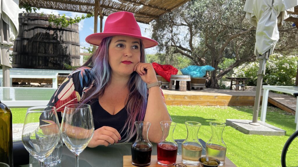 אישה עם שיער ארוך וצבעוני וכובע ורוד בוהק יושבת ליד שולחן חיצוני עם כוסות יין ריקות וקנקנים קטנים של יין, מוקפת בירק ומוצלת על ידי פרגולה מעץ.
