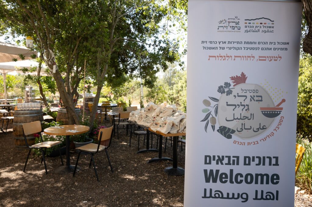 שלט עם כיתוב בעברית, ערבית ואנגלית מקבל את פני המבקרים בפסטיבל. מאחורי השלט, שולחנות וכיסאות חיצוניים מוצבים באזור גן מוצל עם עצים ושקיות על שולחן.