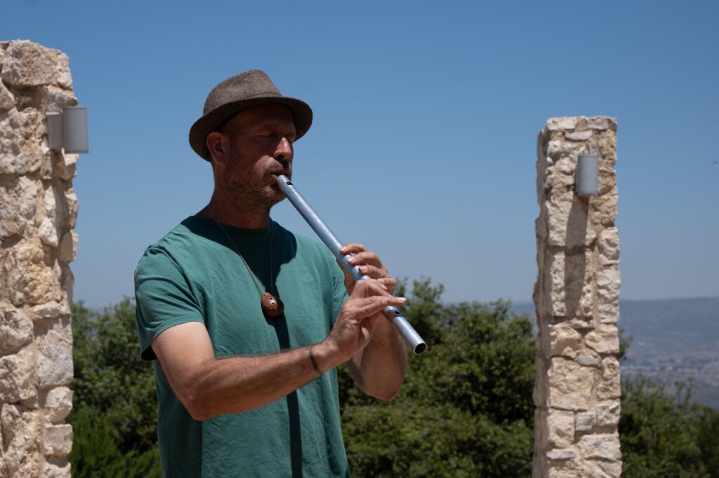 גבר חובש כובע וחולצה ירוקה מנגן בחליל כסף בחוץ, עומד בין שני עמודי אבן כשברקע נראים עצים וגבעות תחת שמיים כחולים וצלולים.