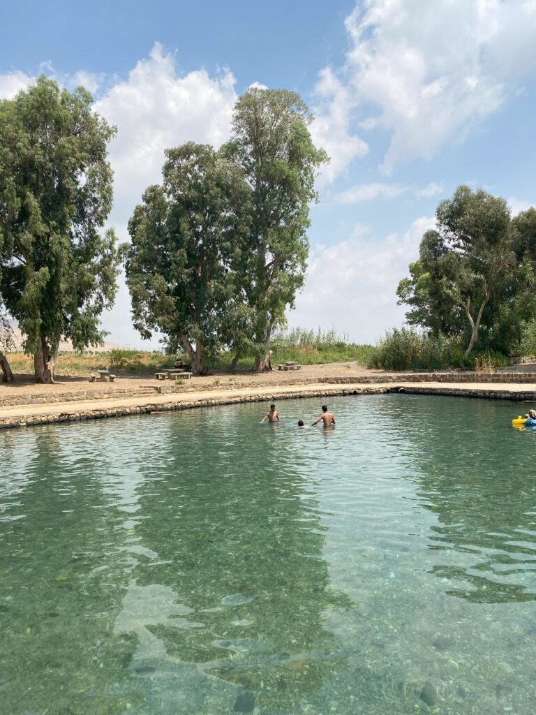 שני אנשים שוחים בבריכה טבעית וצלולה, מוקפים בעצים ירוקים גבוהים וגדות עטויות דשא, תחת שמיים מעוננים חלקית. מצוף צהוב צף בקרבת שפת המים.