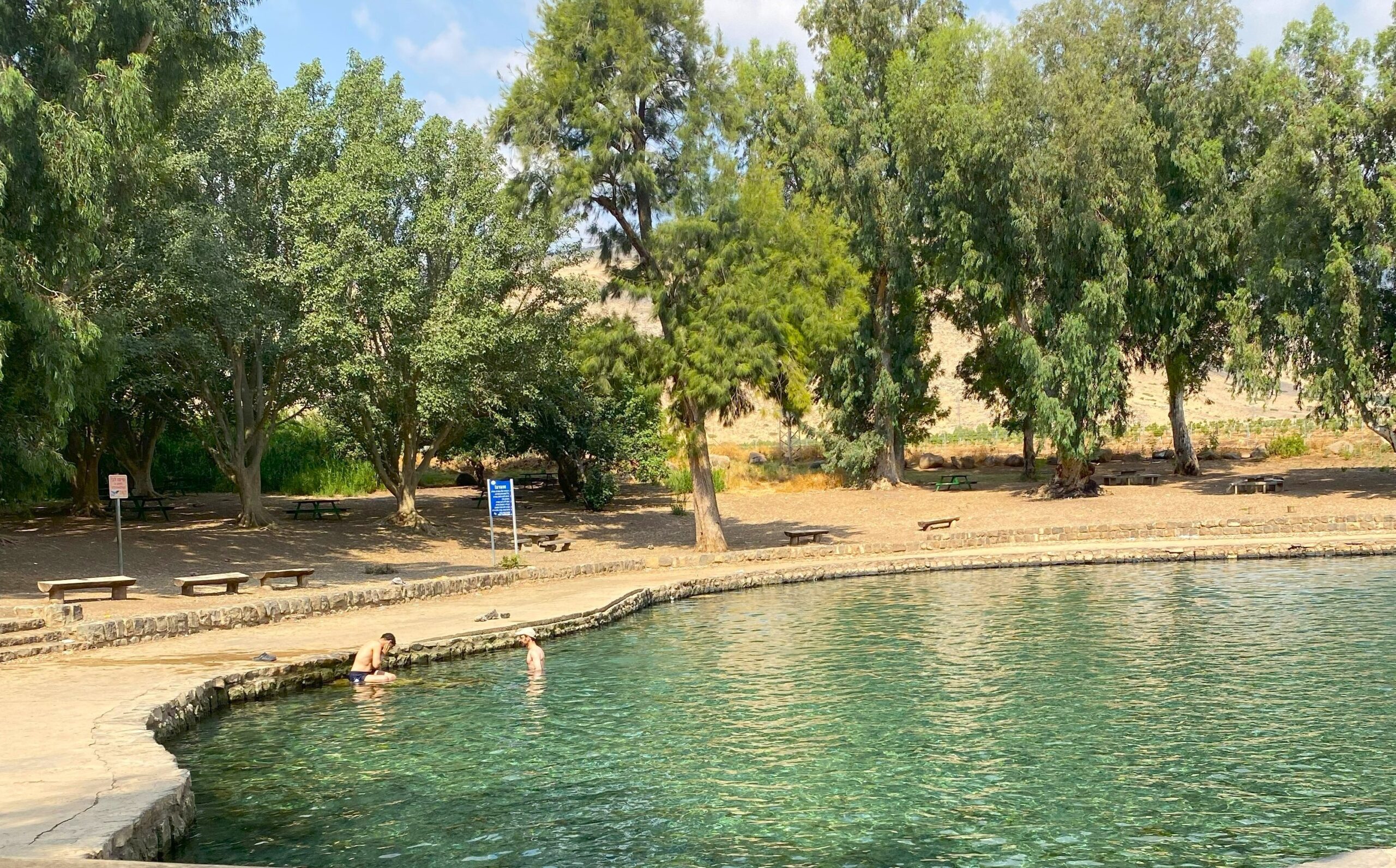 בריכת שחייה טבעית עם מים צלולים מוקפת אבנים, עם שני אנשים במים. ספסלים ועצים גבוהים מקיפים את החוף תחת שמיים מעוננים חלקית.