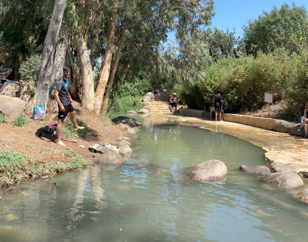 נחל רדוד עם מים צלולים זורם מתחת לעצים, ואנשים משכשכים במים ומנוחים על גדותיו. חלקם יושבים על סלעים או עומדים במים ביום שטוף שמש, מוקפים בצמחייה עבותה.