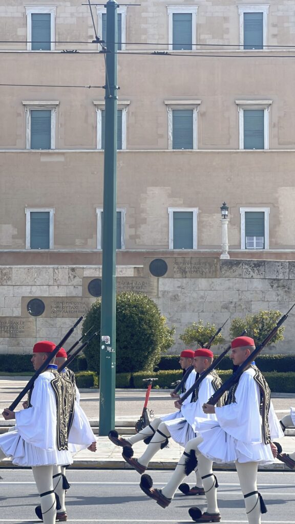 אוזונים, שומרי הנשיא היווני במדים מסורתיים לבנים וכובעים אדומים, צועדים במסדר עם רובים בידיהם מול בניין הפרלמנט היווני באתונה ביום שמש.