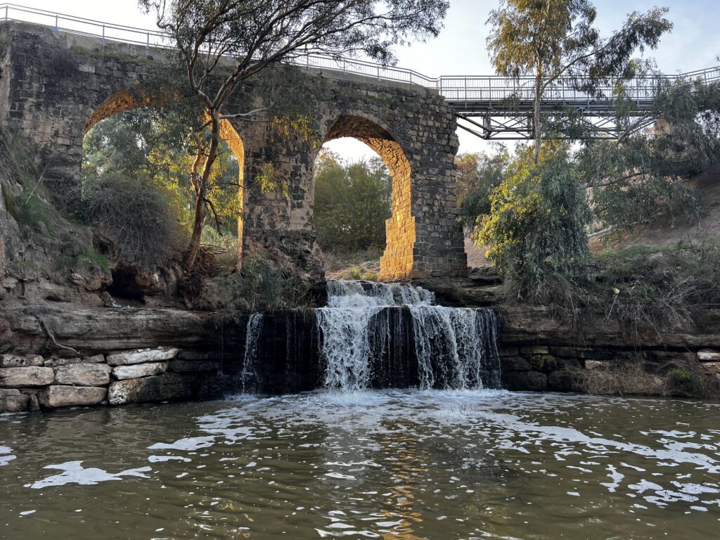 מפל קטן זורם מתחת לגשר קשתות אבן מוקף עצים וצמחייה, כשקרני השמש חודרות מבעד לקשתות ומשתקפות במים שמתחת.