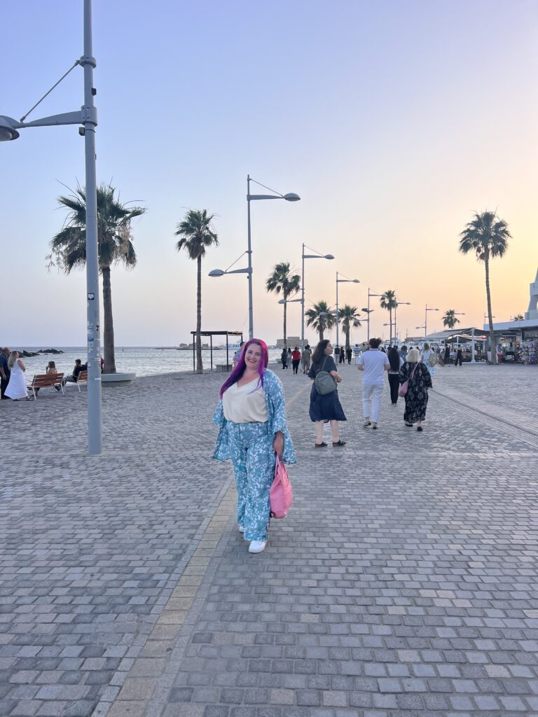 אישה עם שיער ורוד בוהק, לבושה בבגד עם דוגמה כחולה, הולכת לאורך טיילת חוף ים מוקפת דקלים בשעת השקיעה. אנשים אחרים מטיילים בסביבה, וברקע נראים הים והשמים.