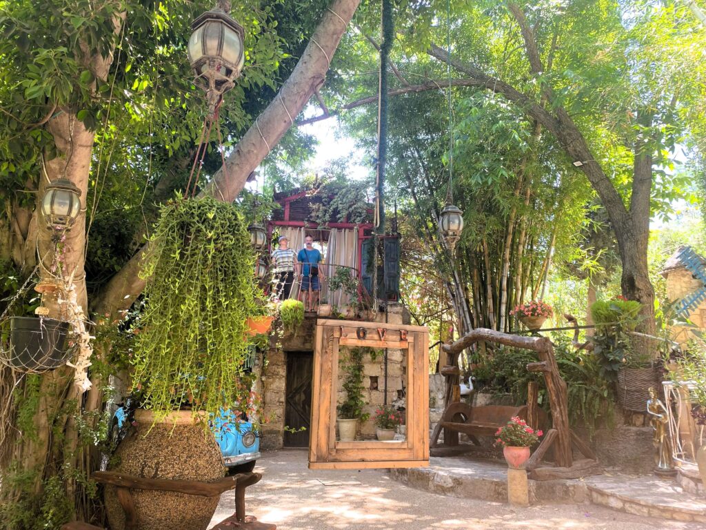 גן שופע ומואר בשמש עם צמחים תלויים, כלי חרס גדולים, נדנדות עץ ופנסים. מבנה אבן כפרי עם מדרגות ואלמנטים צבעוניים ניצב מתחת לעצים גבוהים ועבותים.