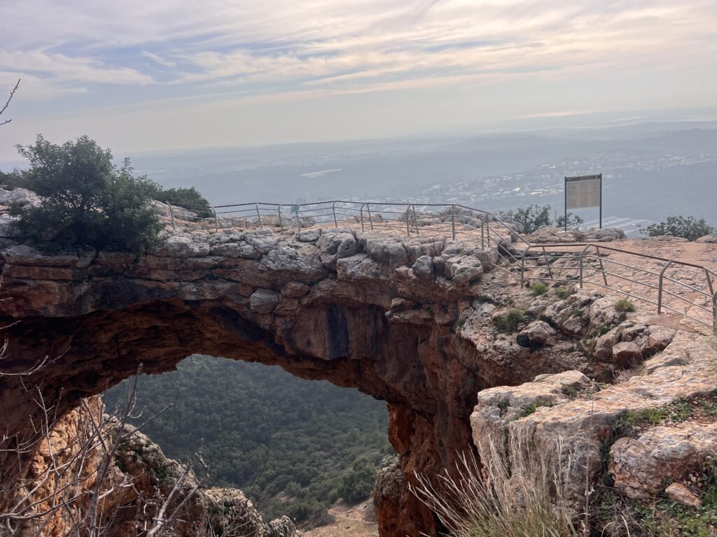 קשת מאבן טבעית עם מעקה מתכת מעליה משקיפה על עמק מיוער וגבעות רחוקות תחת שמיים מעוננים, עם שלט הנראה מימין.