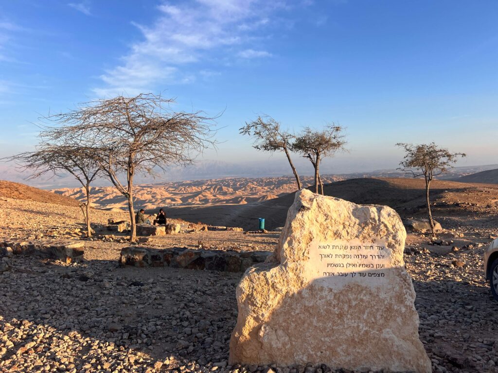 אבן גדולה עם כתובת בעברית ניצבת בנוף מדברי סלעי, עם עצים דלילים וגבעות רחוקות תחת שמים כחולים וצלולים. אדם יושב על ספסל ליד קו האופק.