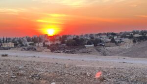 שקיעה כתומה עזה מעל ערד, עם מבנים פזורים מעבר לקדמת התמונה הסלעית והצחיחה. השמש נמוכה, מטילה זוהר חם ומציעה תצפית מדהימה לכיוון ים המלח.