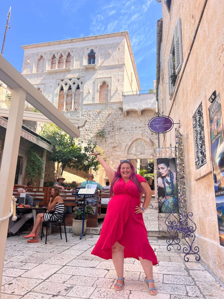 אישה מחייכת בשמלה ורודה בהירה מצטלמת עם יד אחת מורמת בחצר מרוצפת אבן שטופת שמש, מוקפת באנשים היושבים בשולחנות בחוץ ובניינים היסטוריים מאבן בצבע בז'.