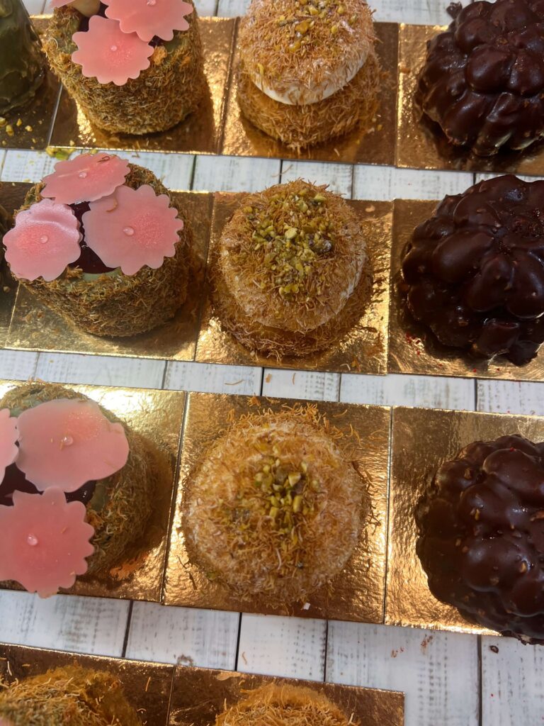 מבחר עוגות מיני אלגנטיות המוגשות על מגשים מוזהבים, עם תוספות כמו קישוטים בצורת פרחים ורודים, פיסטוקים קצוצים וציפוי שוקולד על משטח עץ לבן.