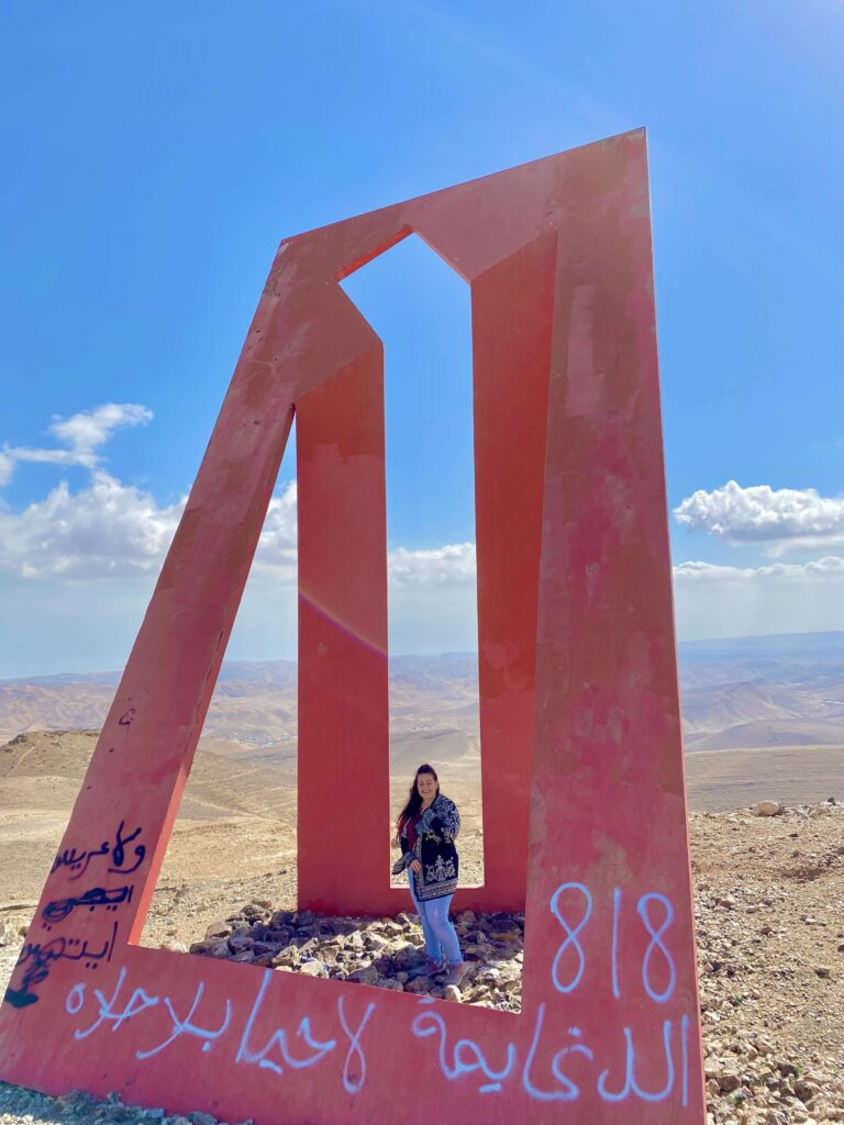אדם עומד בתוך אנדרטה גיאומטרית גדולה בצבע אדום עם צורות חתוכות, הממוקמת על רקע נוף סלעי תחת שמיים כחולים. על האנדרטה נראים גרפיטי בערבית לבנה והמספר 818. ברקע נראים הרים.