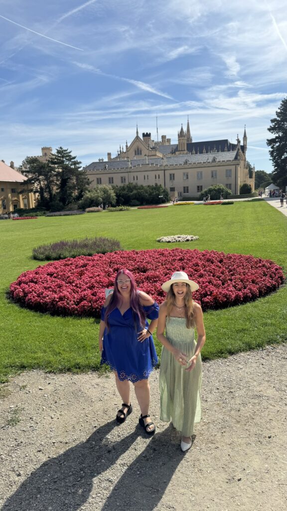 שתי נשים מחייכות ומצטלמות מול ערוגה גדולה עם פרחים ורודים בוהקים, כשברקע מבנה מפואר ומקושט ומדשאות ירוקות תחת שמיים כחולים ושטופי שמש.