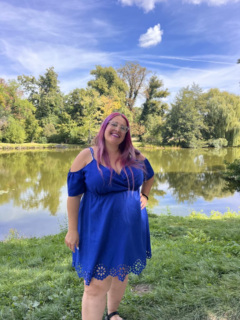 אישה עם שיער ורוד ארוך, לבושה בשמלה כחולה בהירה, עומדת מחייכת מול בריכה מוקפת בעצים ירוקים תחת שמיים כחולים עם עננים דקים.