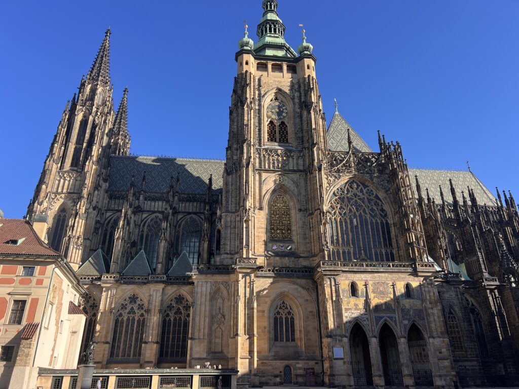 קתדרלה גותית גדולה עם צריחים גבוהים, קשתות מחודדות ועבודת אבן מפורטת ניצבת תחת שמיים כחולים וצלולים. אור השמש מדגיש את חזיתה המורכבת וחלונות הוויטראז' שלה.