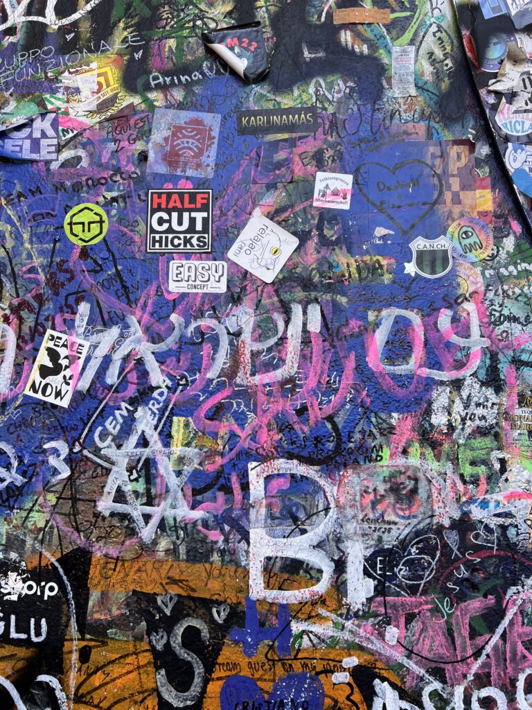 קיר צבעוני מכוסה גרפיטי, מדבקות וציורים, כולל טקסט שרבוטים, תגיות וסמלים בצבעים שונים. מדבקות בולטות כוללות HALF CUT HICKS ו-EASY. טוש לבן נראה בחלק העליון.