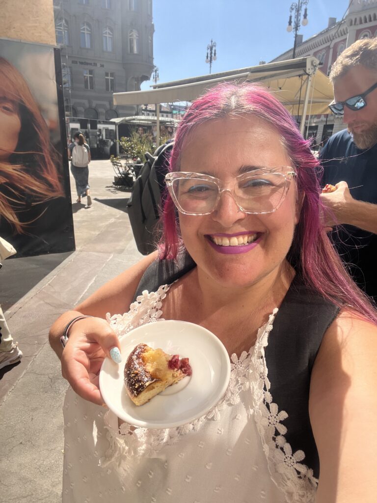 אישה מחייכת עם שיער ורוד ומשקפיים שקופים מחזיקה צלחת עם מאפה בחוץ, בסביבה עירונית שטופת שמש. ברקע נראים אנשים ובניינים.