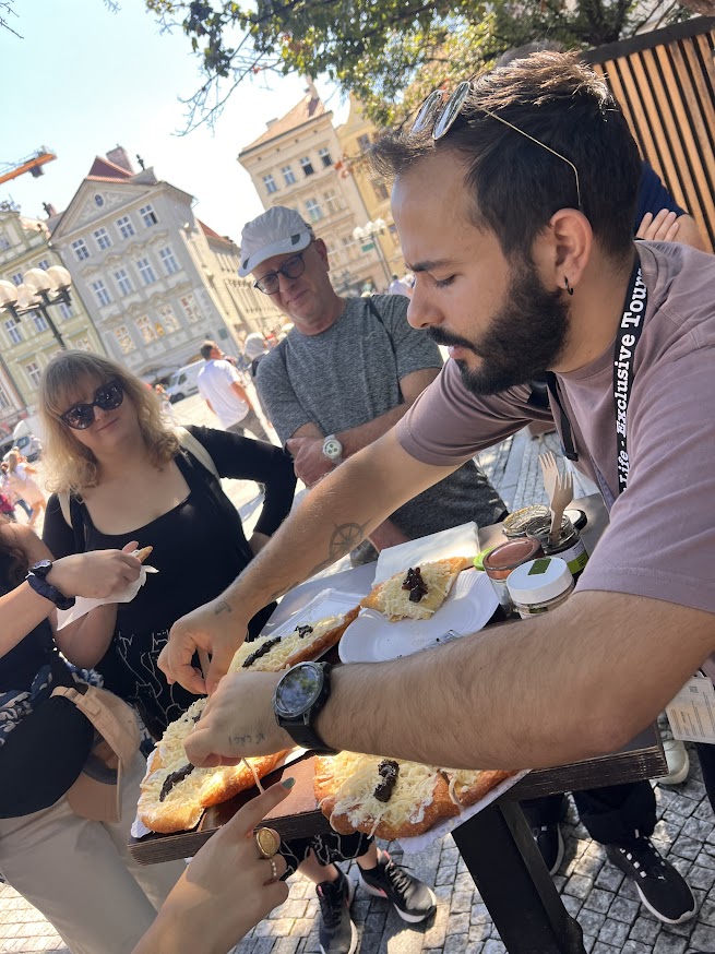 גבר מגיש מאפים עם שמנת ופירות לקבוצת אנשים בכיכר עירונית אירופית, כשברקע נראים מבנים היסטוריים.