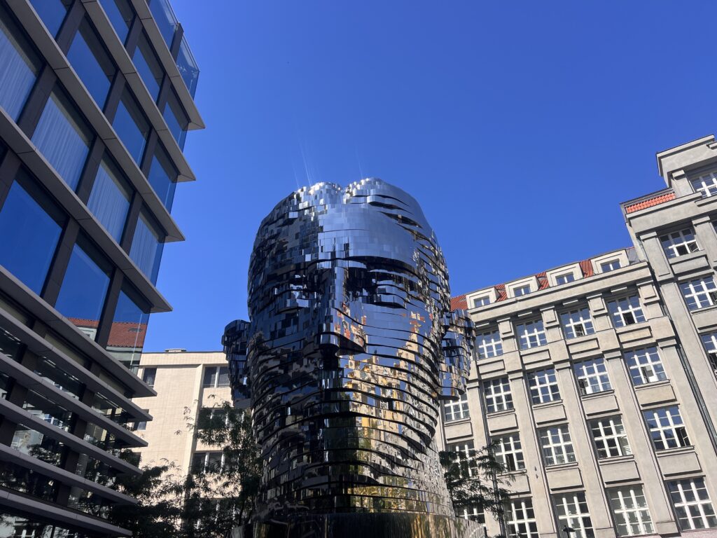 פסל מראה גדול של ראש אדם, המורכב משכבות אופקיות מסתובבות, ניצב בין מבנים מודרניים והיסטוריים תחת שמיים כחולים וצלולים.