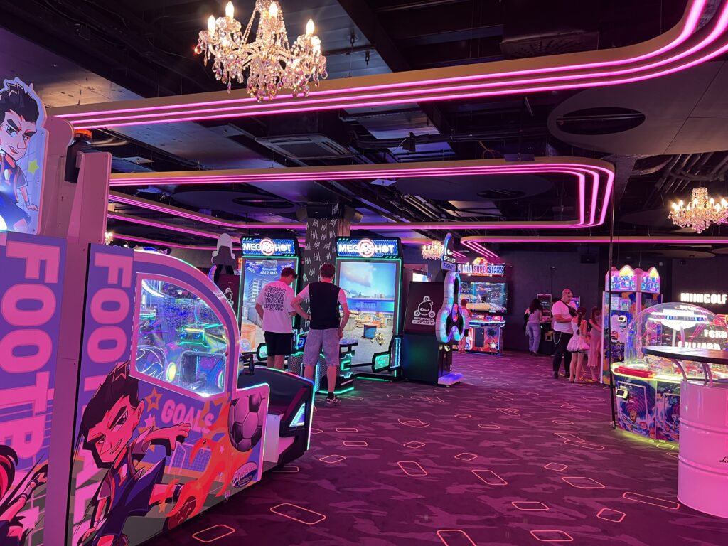 אולם משחקים מואר בנורות ניאון, שבו אנשים משחקים במכונות משחק צבעוניות ומגוונות. נברשות תלויות מהתקרה, והאווירה תוססת עם אורות בוהקים ומסכי משחק הפזורים ברחבי החדר.