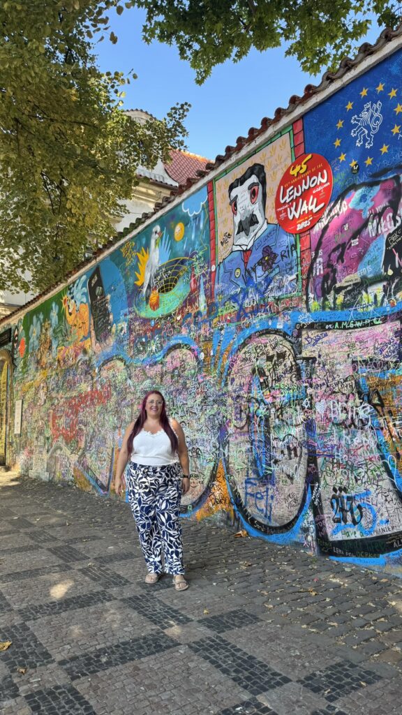 אישה עם שיער ארוך וכהה, לבושה בחולצה לבנה ומכנסיים מעוצבים, עומדת מול קיר צבעוני מכוסה גרפיטי עם ציורים וטקסטים שונים, כולל שלט שעליו כתוב "קיר לנון", מתחת לעצים עלים.