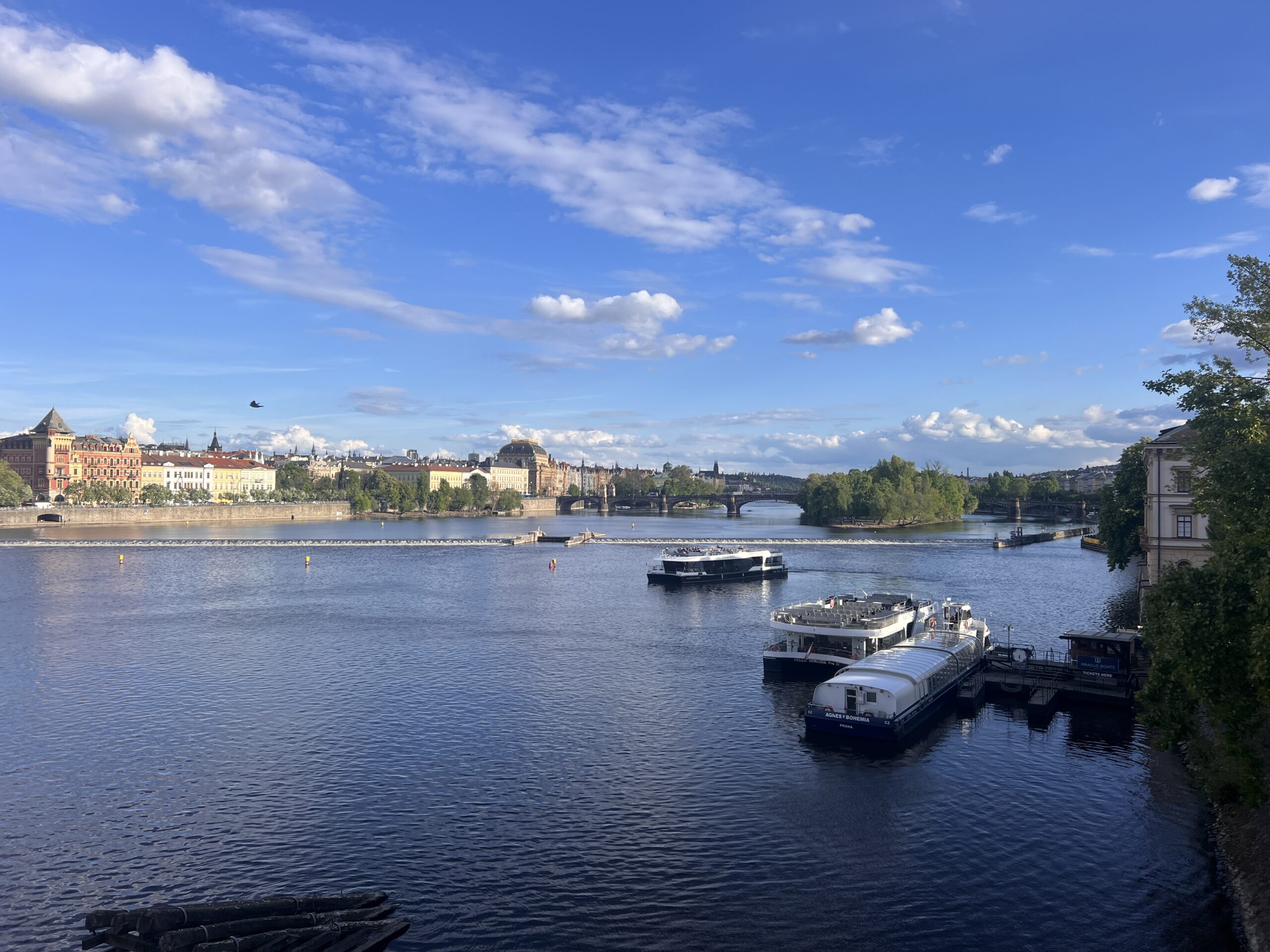 נהר רחב עם מספר סירות עוגנות ועצים ירוקים ושופעים על גדותיו, תחת שמיים כחולים עם עננים פזורים. מבנים וגשרים נראים מרחוק.