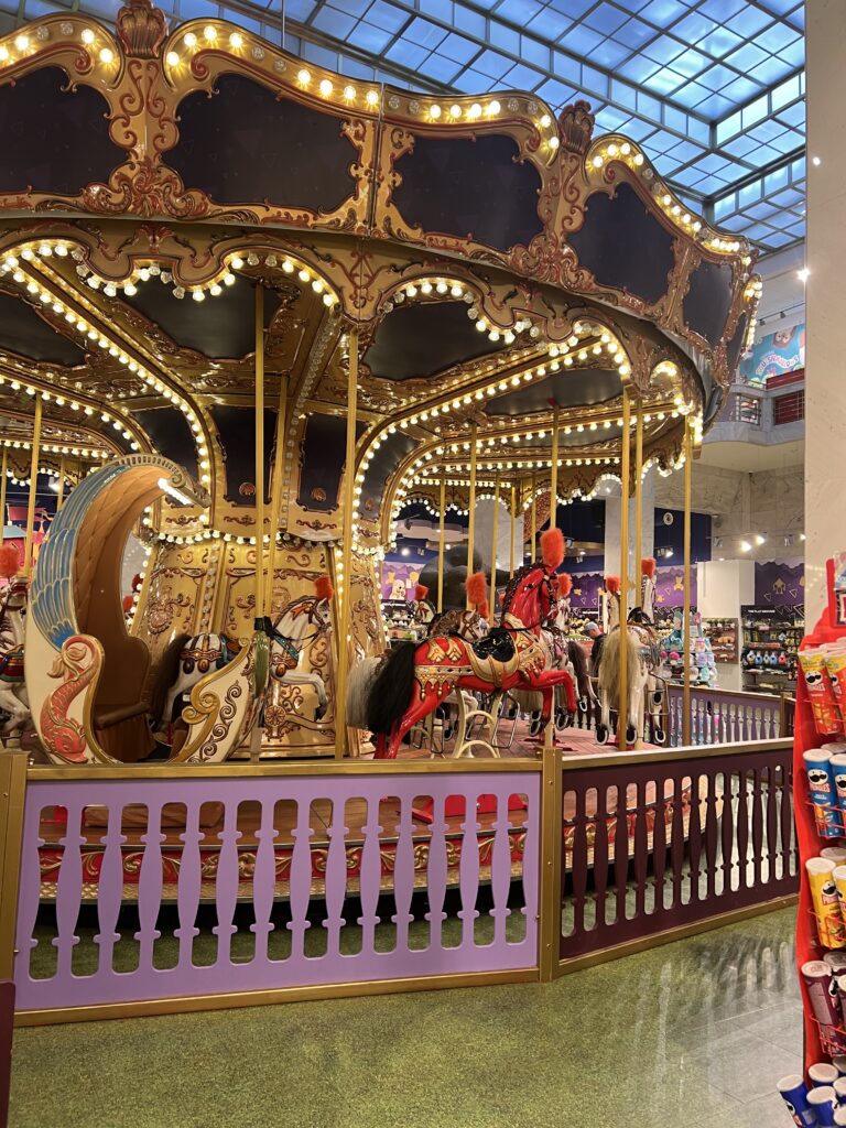 קרוסלה מקורה ומוארת, עם סוסים וכרכרות מעוטרים, ניצבת מאחורי גדר סגולה ובורדו, מוקפת בתצוגות צבעוניות ומתחת לתקרת זכוכית.