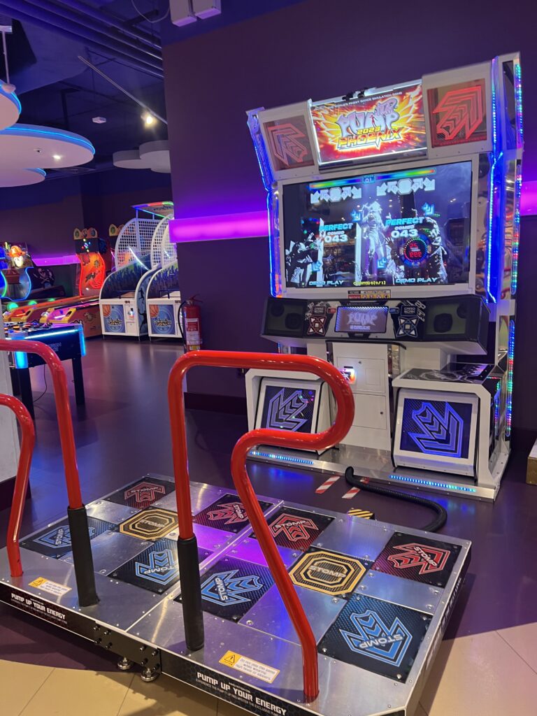 מכונת ארקייד Dance Dance Revolution עם פלטפורמת ריקוד מתכתית ומעקות אדומים ניצבת באולם משחקים מואר בנורות ניאון, מוקפת במכונות משחק צבעוניות אחרות.