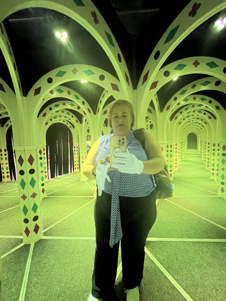 אדם לבוש בחולצה כחולה עם נקודות, מכנסיים שחורים וכפפות לבנות מצלם סלפי במבוך מראות עם קשתות ירוקות וצהובות ודוגמאות בצורת יהלום, היוצרות השתקפויות מרובות.